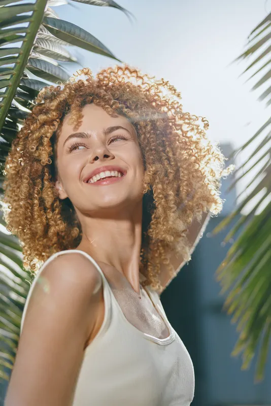 smiling woman with curly hair enjoying sunlight outdoors, bright joyful expression, tropical leaves background, summer vibe, natural beauty radiance