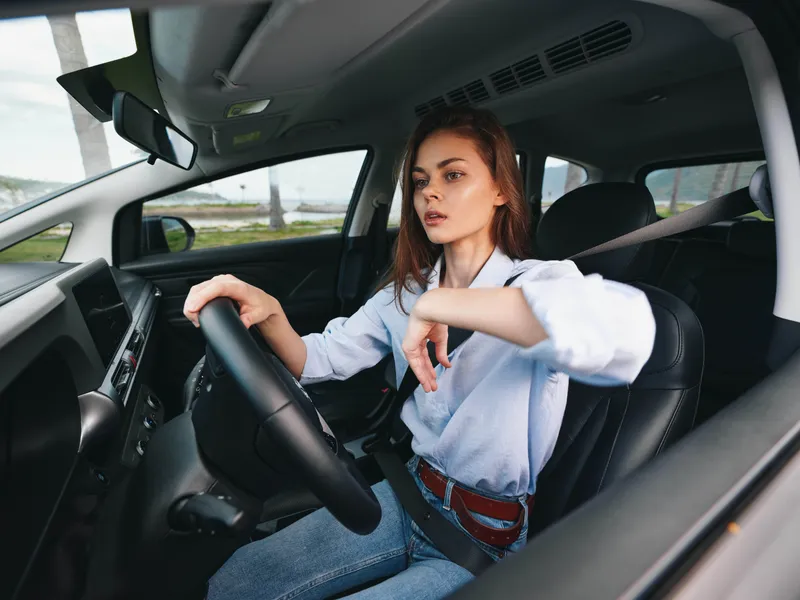 Женщина на водительском сиденье автомобиля, положив руку на руль, смотрит прямо в камеру, излучая уверенность и сосредоточенность