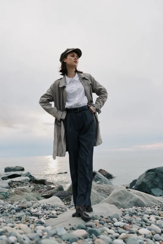 Confident young woman stands on rocky beach at sunset wearing casual jacket, cap, and trousers. Moody sky and calm sea create peaceful outdoor atmosphere and fashion concept.