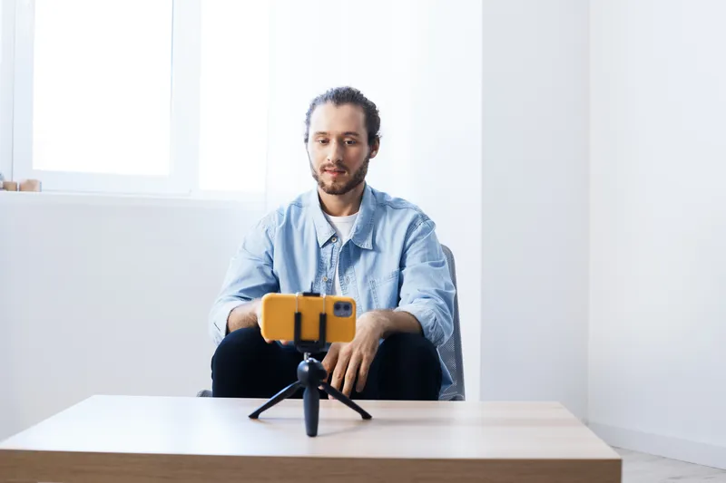 Young man filming a video with a smartphone on a tripod, feeling focused and relaxed in a bright, modern room, ideal for creative content and digital communication