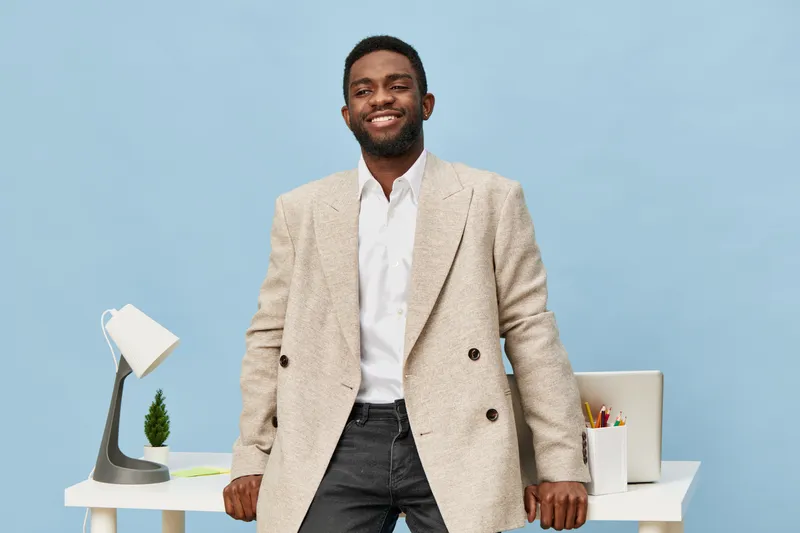 Confident smiling man with dark skin wearing beige blazer and white shirt standing in modern office background. Business professional youthful style, casual elegance, workspace environment, confident 