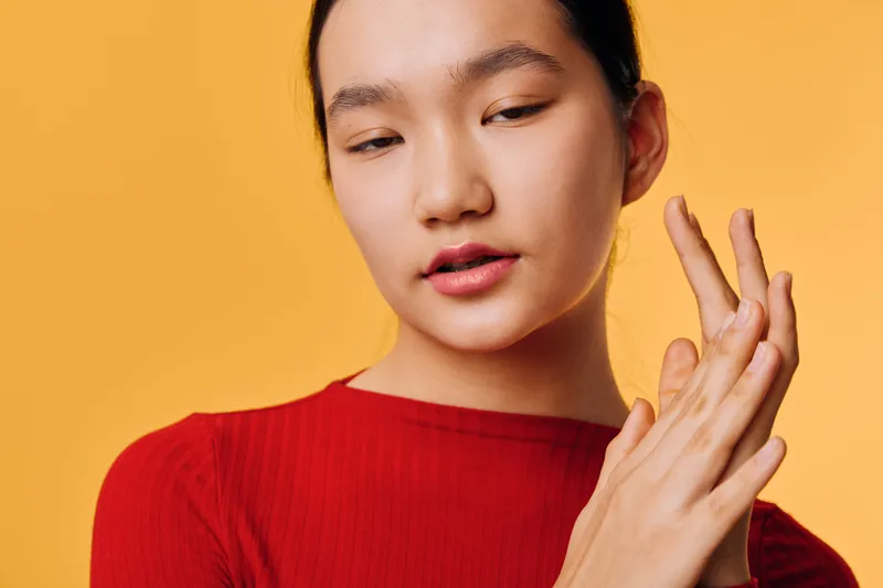 Young woman with smooth skin wearing red top posing against yellow background with eyes closed and hands touching face in a skincare or beauty concept portrait.