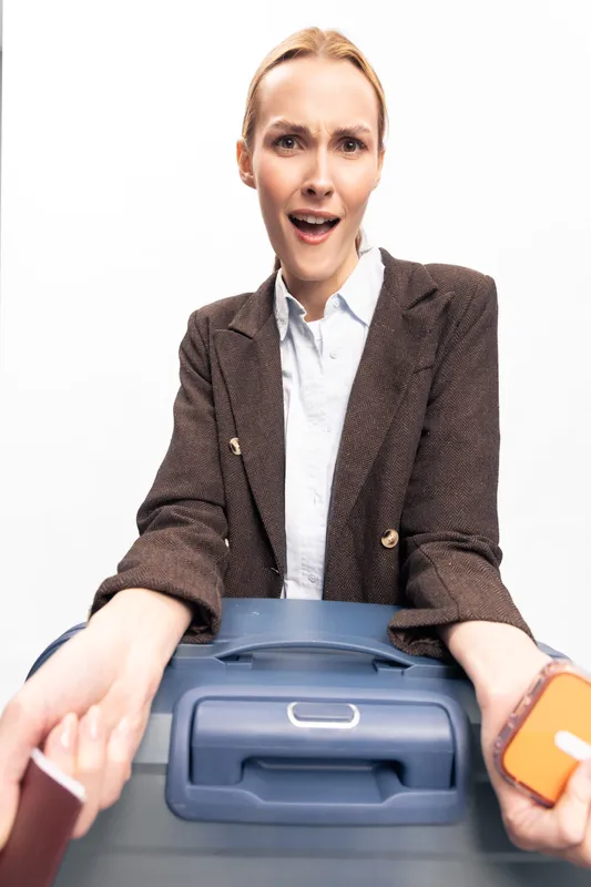 Surprised woman in brown jacket holding blue suitcase and passport with boarding pass, expressing shock or confusion. Concept of travel, airport, and unexpected situations.