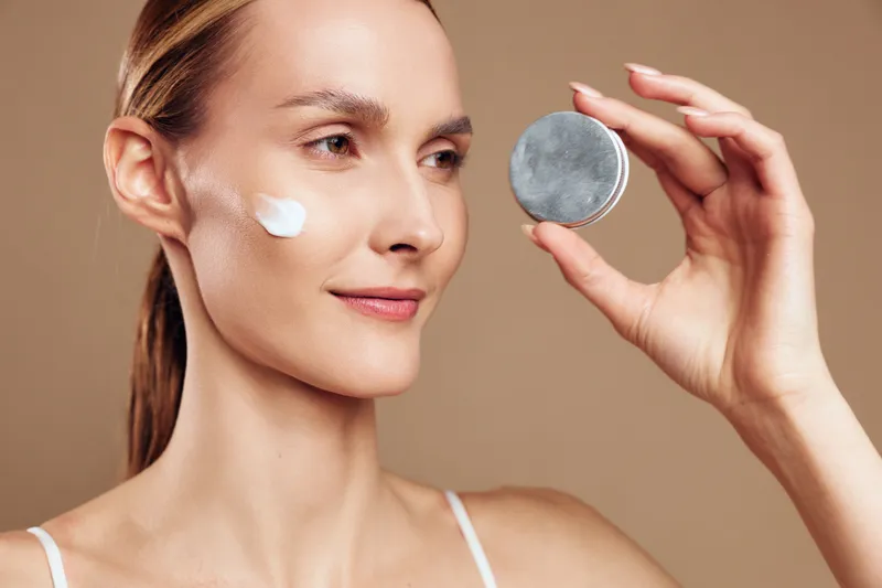 Young woman applying face cream with smooth skin, natural makeup, holding skincare container, beige background, beauty routine, moisturizing effect, healthy lifestyle concept