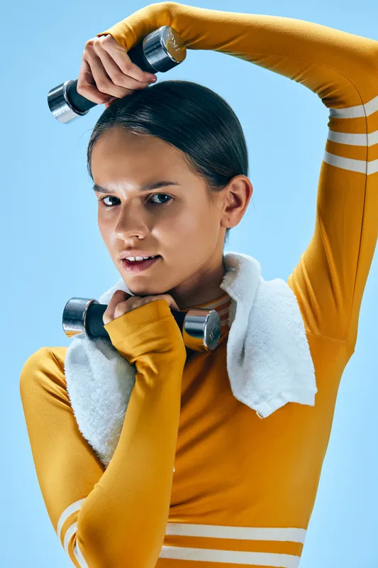 Woman fitness athlete performing upper body workout with dumbbells, wearing a yellow athletic top and towel around the neck, posed against a blue studio backdrop.