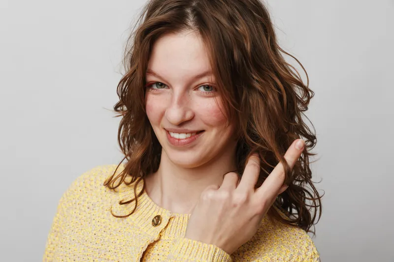 Smiling woman with curly hair wearing yellow sweater shows middle finger gesture. Studio portrait on grey background. Concept of attitude, defiance, and casual expression in young adult.