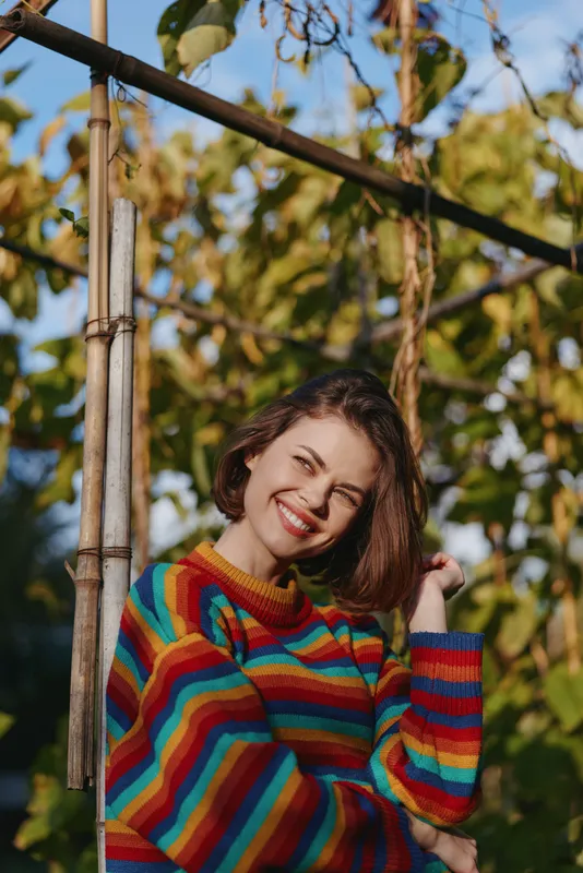 Woman smile sweater rainbow portrait outdoors happy nature: Young woman in a colorful striped sweater smiling brightly in a sunlit garden, short hair framing face and green foliage background.