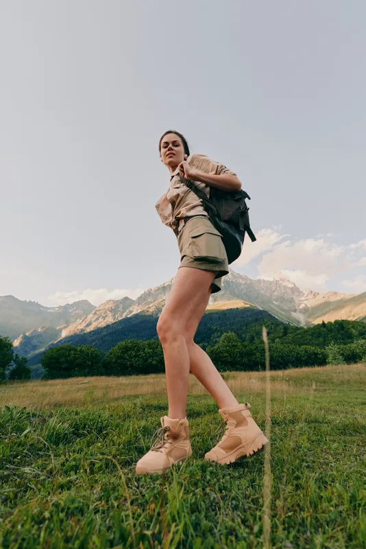 Active woman hiking through a field with a backpack, mountains in the distance, and clear sky. Outdoor lifestyle scene focused on exploration, nature, and travel moments.