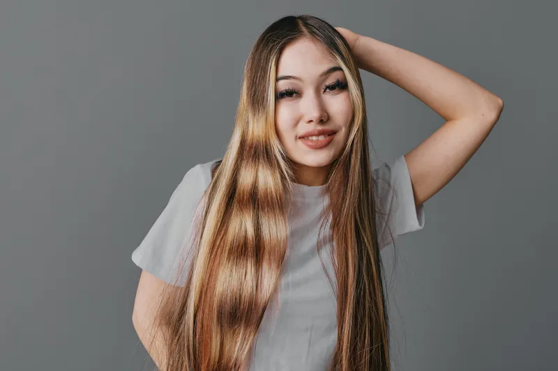 Young woman with long straight hair posing against gray background smiling and looking at camera in casual white t shirt with hand behind head.
