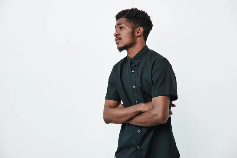 Young black man with dark skin wearing black shirt standing with arms crossed looking sideways on white background. Portrait of confident African ethnicity male in casual style.
