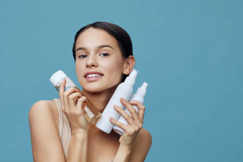 Young woman holding skincare products with a joyful expression against a soft blue background, showcasing beauty and healthy skincare concepts