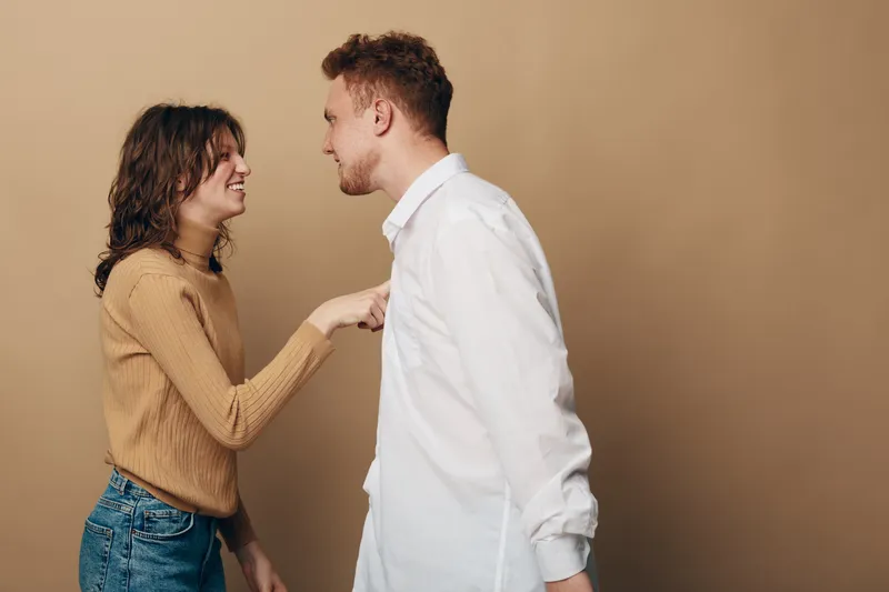 A young woman in a brown sweater playfully points at a man in a white shirt. Both smile and engage in a lighthearted conversation against a beige background.