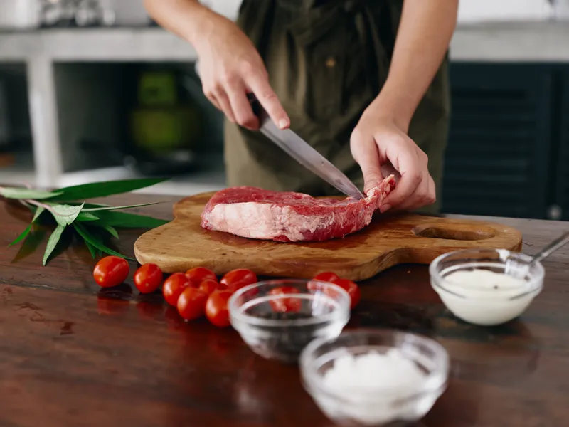 Женщина с ножом в руке нарезает свежее мясо стейка для жарки на кухне