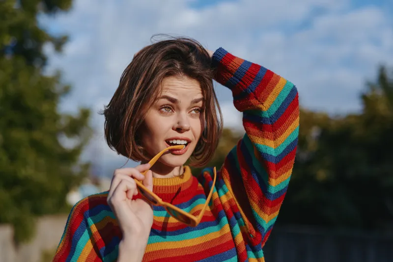 Woman portrait in colorful striped sweater holding sunglasses outdoors, thoughtful expression and casual fashion style, natural light, shallow depth of field, warm autumn tones.