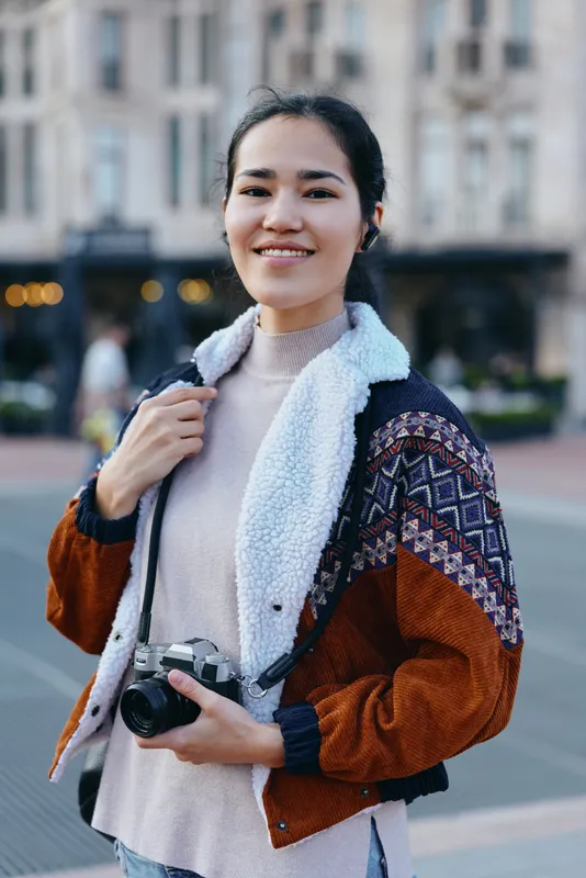 Woman with camera on street in casual attire, holding a warm jacket and soft sweater, ready for street photography and everyday moments, urban backdrop, candid expression, creative lens focus,