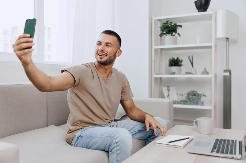 Happy man taking a selfie in a modern living room, wearing casual clothes and smiling while relaxing on a comfortable sofa. Perfect for lifestyle and technology themes.