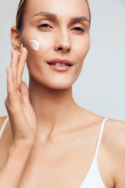 Young woman with clear skin applying white face cream on cheek. Close-up portrait with natural light. Concept of skincare, beauty routine, moisturizing, and healthy lifestyle.