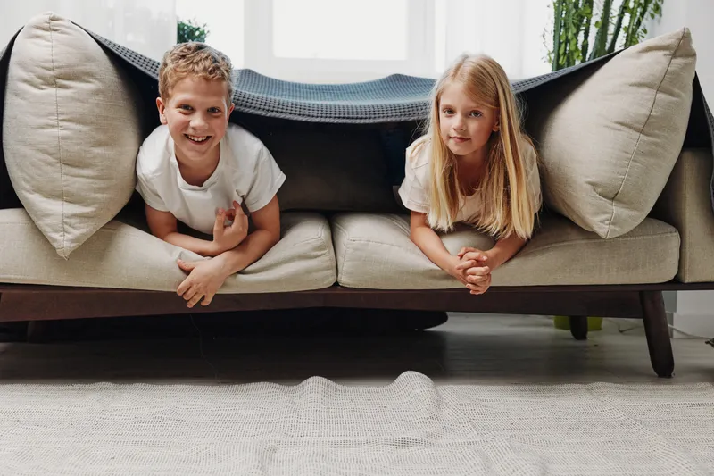 Two cheerful children lying under a blanket on a modern sofa in a bright living room. Concept of playful moments, family bonding, and cozy home environment for kids.