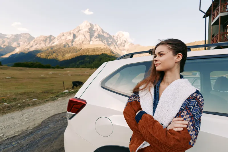Woman and car parked in countryside by mountains for travel and roadtrip, standing in sweater jacket with folded arms, scenic landscape, outdoor adventure and calm moment.