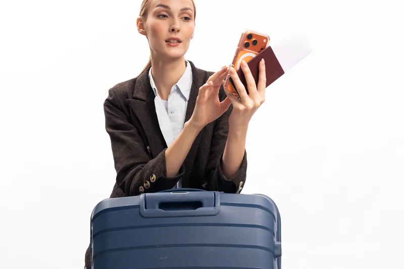 Young woman in business attire holding passport and smartphone behind large suitcase on white background. Concept of travel preparation, business trip, and modern tourism technology.