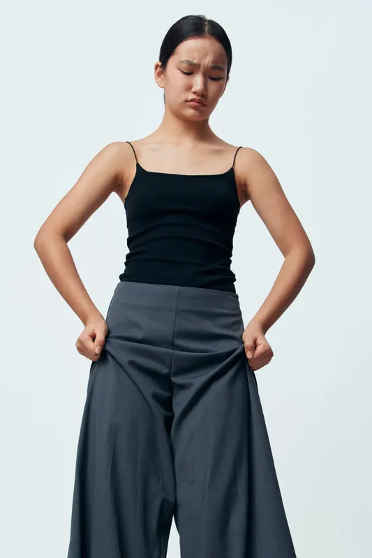 Young woman trying on oversized wide gray pants with worried expression, hands inside pockets, wearing black tank top against white background in studio. Clothing, fashion, size, style.
