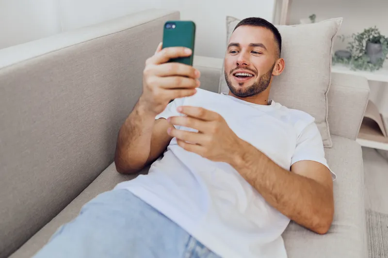 Happy man relaxing on a sofa, engaging with his smartphone, showcasing modern lifestyle and technology trends for 2025. Perfect for social media content.