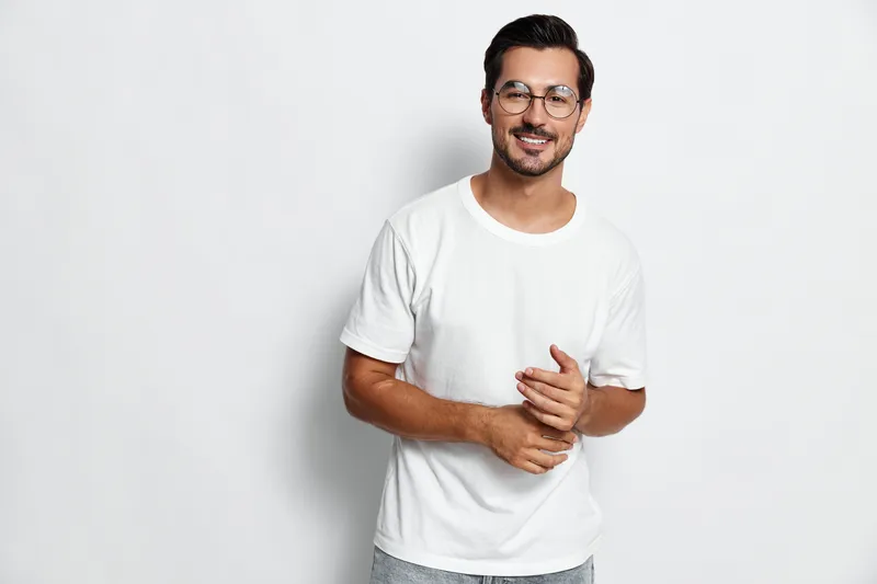 Smiling man in casual white t-shirt with glasses stands confidently against plain background while posing with friendly expression. People lifestyle concept.