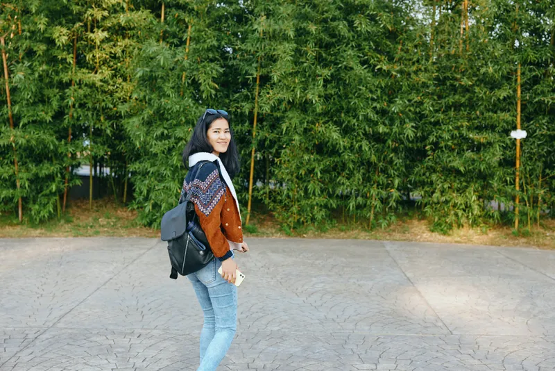 Woman with backpack walking on a paved path beside green trees, casual style, cheerful pose, outdoor lifestyle and travel vibe for everyday moments
