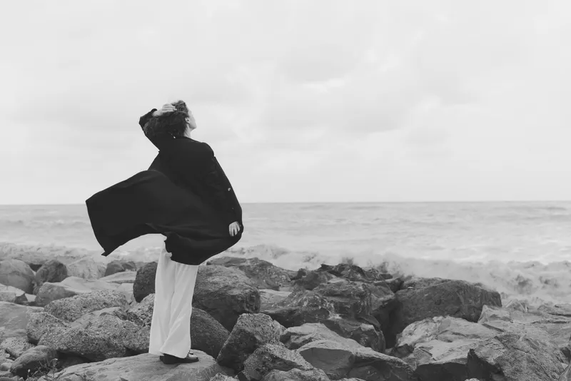 Windswept cloak flows as solitary figure stands on rocky shore, facing the sea, capturing movement, silence, and rugged coastline in monochrome