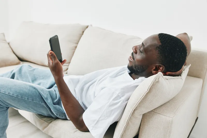 Relaxed young Black man on a sofa, reading on his smartphone, casual style, comfortable home environment, soft lighting for a cozy atmosphere