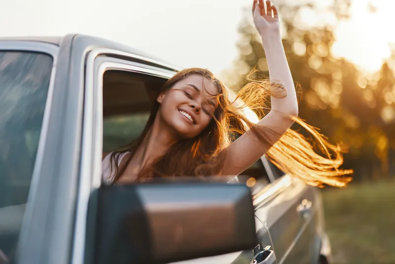 Young woman enjoying a road trip with her hair flowing in the wind, smiling happily from a car window on a sunny day, capturing the essence of freedom and adventure.