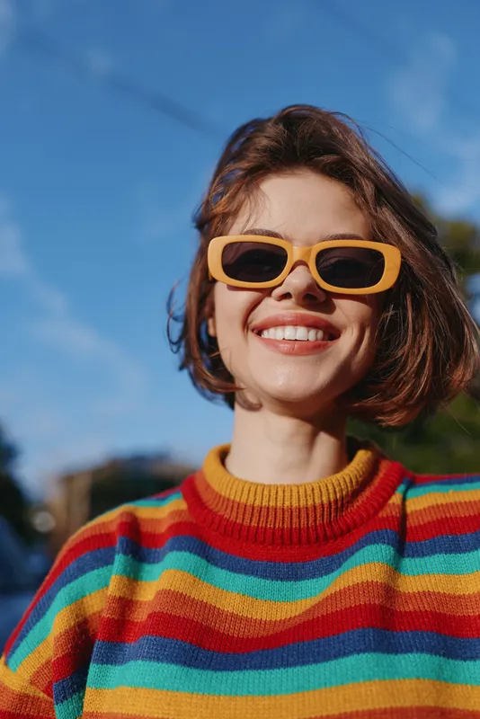 Woman smiling in yellow sunglasses and colorful sweater portrait outdoors, happy fashion closeup with short hair, sunlight and blue sky for lifestyle and casual seasonal style.