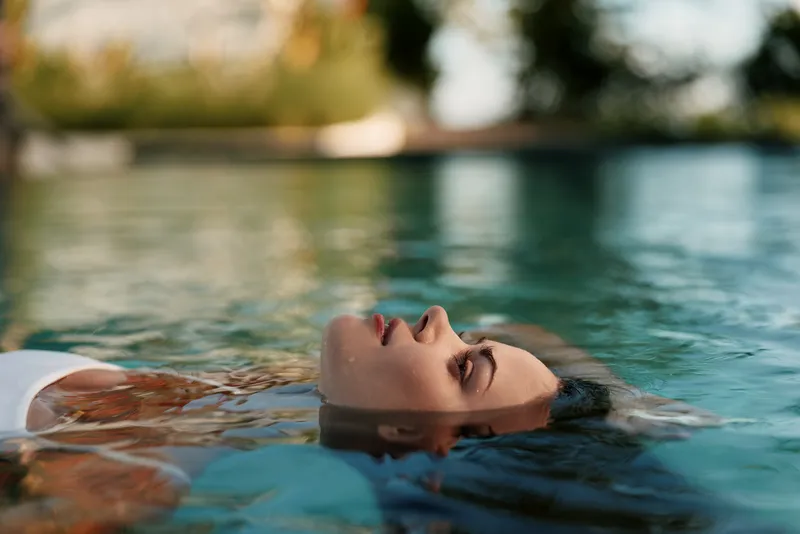 Pool relaxation, woman floating on her back with eyes closed, white swimsuit, peaceful mood. Sunlit water and distant greenery create a tranquil moment focused on lifestyle and wellness.