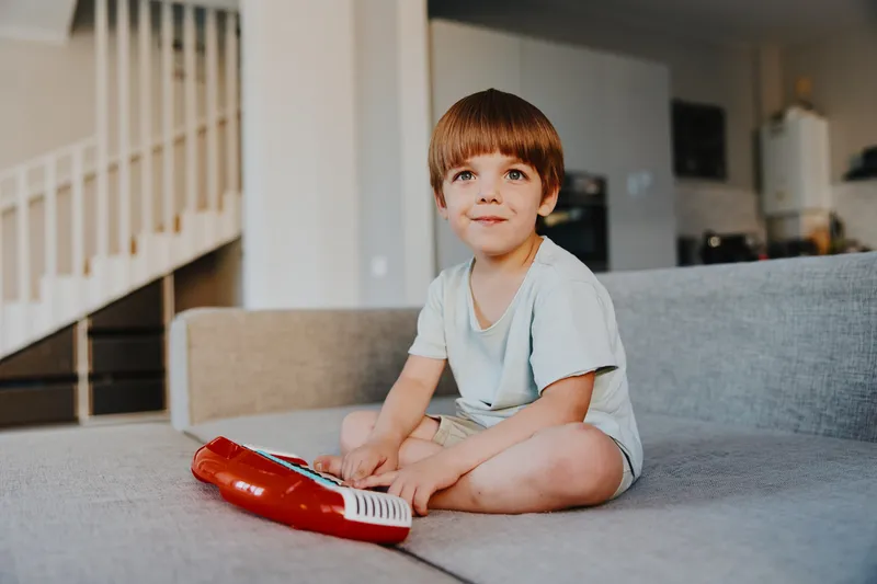 Happy boy playing with a toy keyboard, engaging in music while sitting on a soft couch in a cozy living room, showcasing childhood joy and creativity.