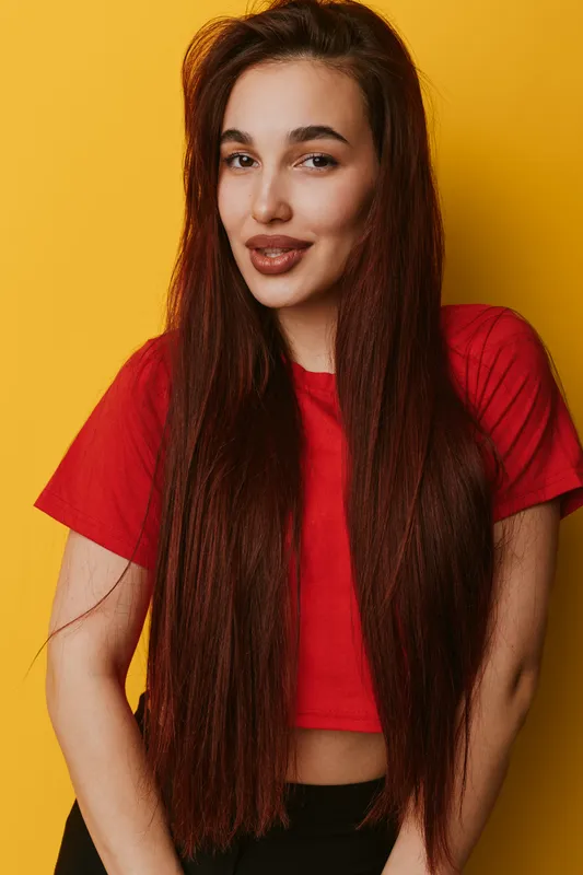 Woman with long straight hair wearing red crop top posing against yellow background smiling confidently. Portrait of young female with natural makeup and casual style in studio.