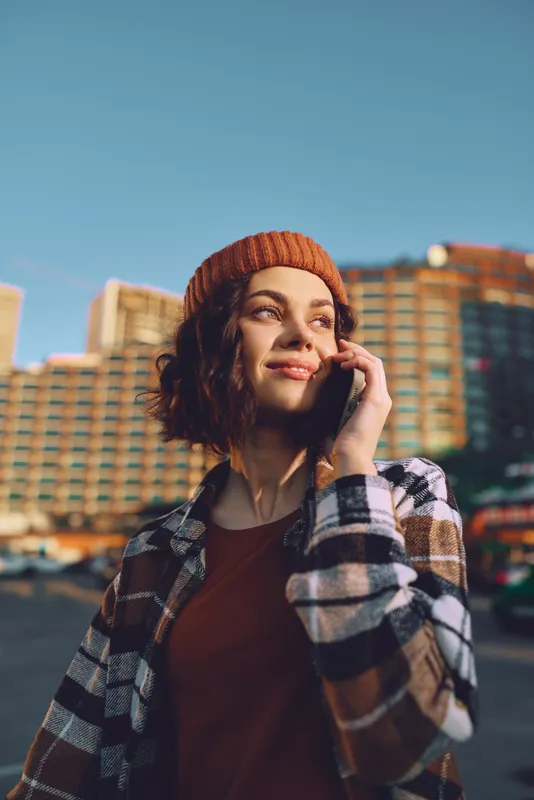 Woman on phone in urban streets, beanie and plaid jacket, smile in golden light, candid lifestyle portrait conveying authenticity, mindful living and emotional storytelling with warm glow.