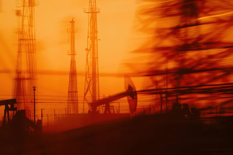 Силуэты нефтяных насосов и телекоммуникационных вышек сливаются с теплыми оранжевыми оттенками заката, подчеркивая энергетическое производство и технологии