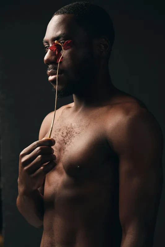 Black man with dark skin holding dried flower near face in creative conceptual visual style. Artistic portrait with bare chest and thoughtful expression on dark background.