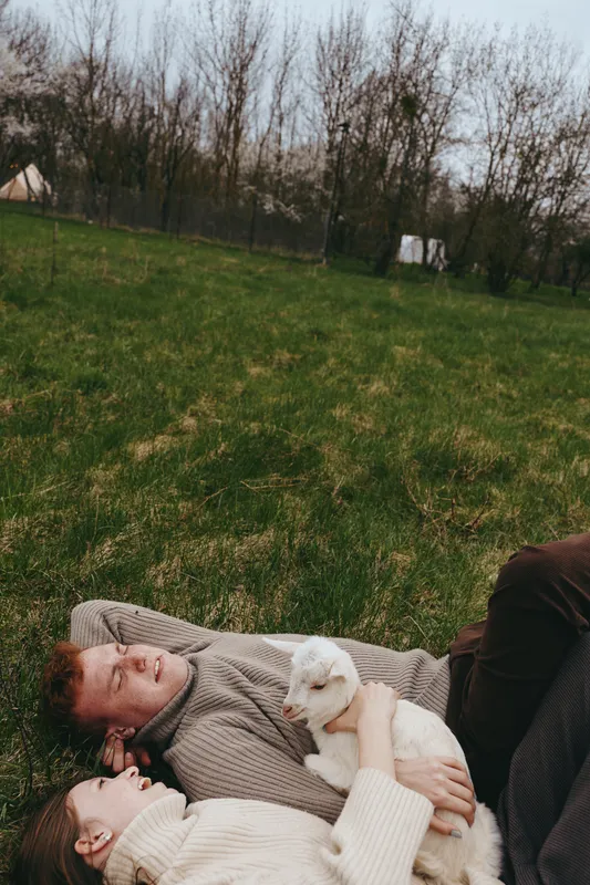 Relaxed couple outdoors on a grassy field cuddle a white goat kid, cozy sweaters, natural light, intimate lifestyle moment, tranquil rural setting, warm affection and companionship