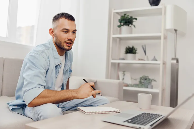 Focused man working remotely in a bright and cozy living room, wearing casual clothes and taking notes. Ideal for illustrating modern work-life balance concepts.