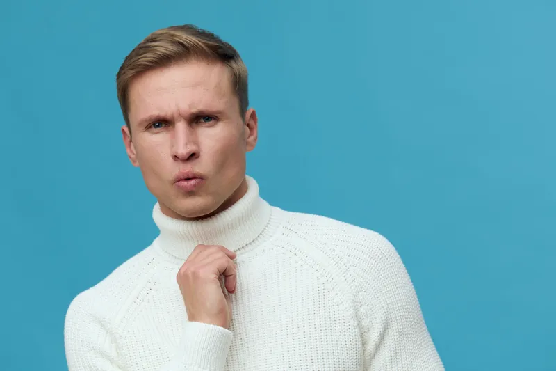 Close-up of confused man in white turtleneck sweater against blue background, studio portrait, human facial expression, emotion, fashion look, indoor setting.