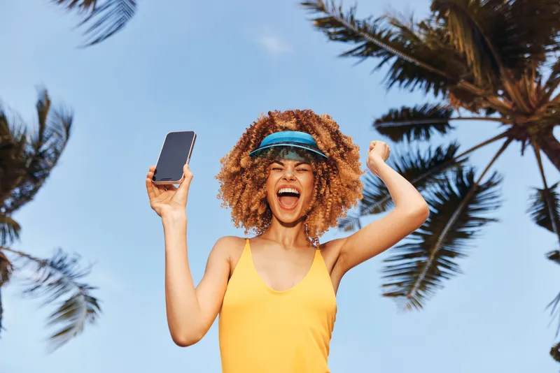 Весёлая девушка в жёлтом купальнике и козырьке держит смартфон на фоне ясного голубого неба с пальмами