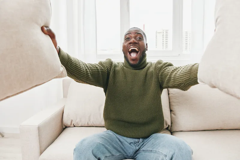 Joyful black man celebrating with pillows in a modern living room, showcasing happiness and comfort in a cozy setting, perfect for lifestyle and home concepts