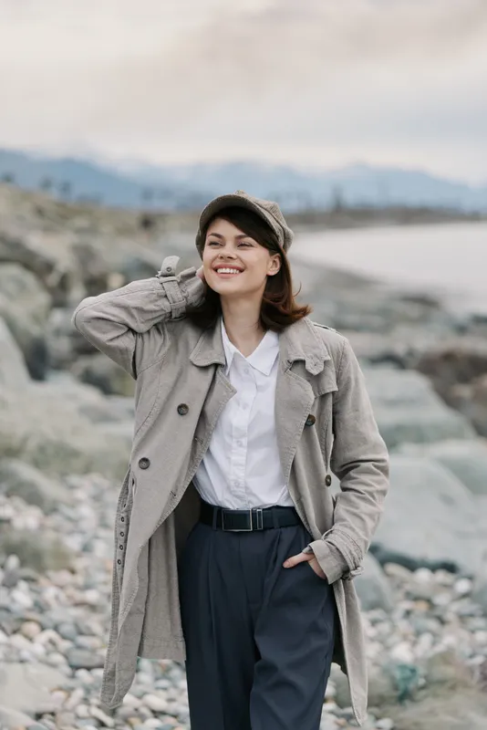 Smiling woman in a textured coat and cap stands on a rocky beach, hand behind head, looking away. Casual autumn outfit with relaxed mood and natural coastal background in soft light.