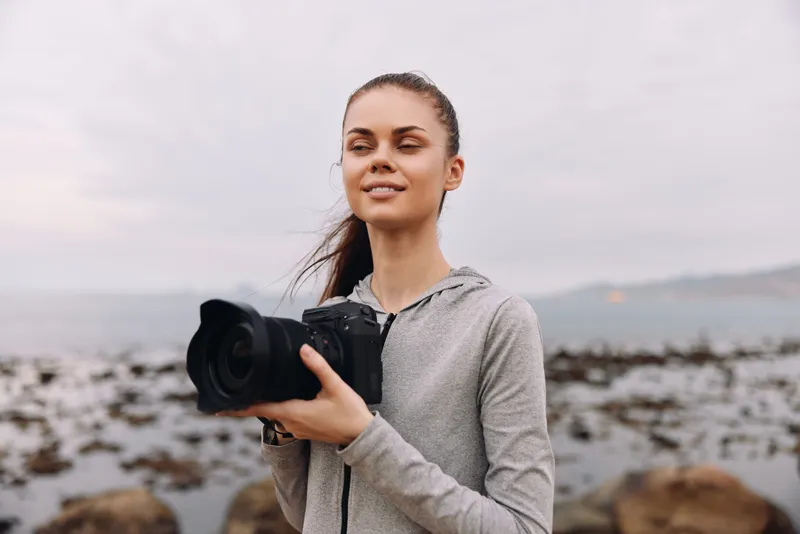 Young woman with a camera on a beach during sunset, showcasing confidence and passion for photography, wearing a casual gray hoodie, with a scenic ocean background