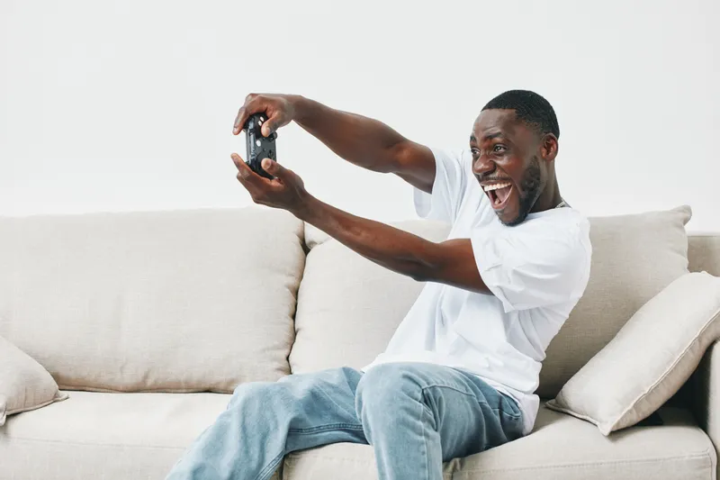 Happy young African man enjoying video games on a couch, showcasing excitement in a casual home setting, perfect for gaming niche