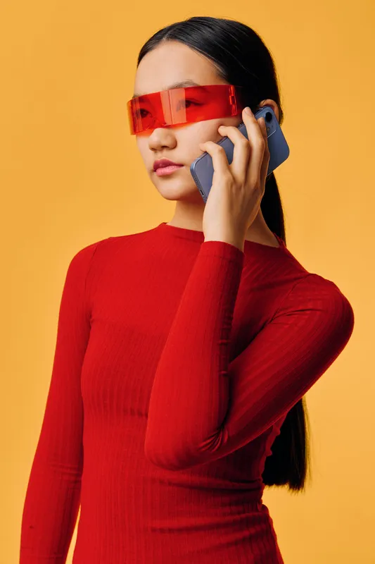 Young woman in red long sleeve top and futuristic red glasses talking on smartphone against orange background. Stylish female portrait with sleek hairstyle and modern accessories.
