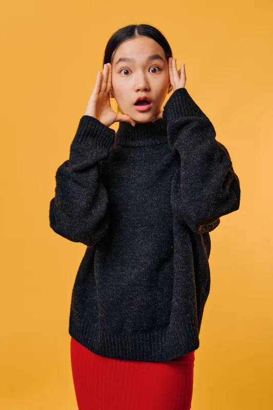 Surprised young woman with open mouth holding hands on head wearing knitted black sweater and red skirt standing on orange background indoors. Emotion, gesture, astonishment expression.