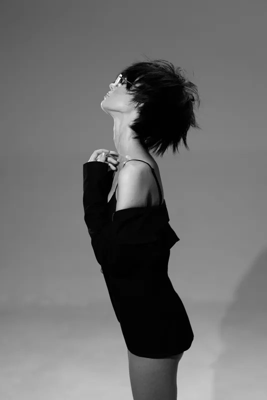 Stylish woman in black dress posing elegantly with eyes closed and short hair in black and white studio setting.