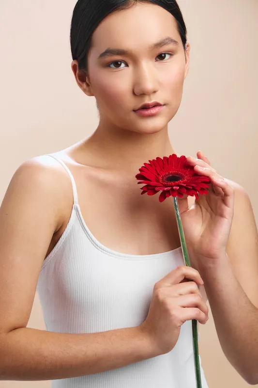Asian woman holds a red gerbera flower in front of a neutral studio backdrop. She wears a white tank, gazing softly at the camera, exuding calm elegance and gentle confidence.